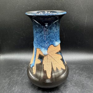May include: A ceramic vase featuring a flared rim and a bulbous base. The upper portion is glazed in a mottled blue with drip effects, while the lower section displays a natural tan clay finish with a prominent embossed maple leaf design.