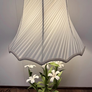 Peut inclure: Une lampe de table avec un abat-jour en tissu plissé blanc à bord festonné et bordure grise. La base représente un bouquet de lys blancs avec des tiges et des feuilles vertes, fixé sur un socle carré en pierre blanche et bois foncé.