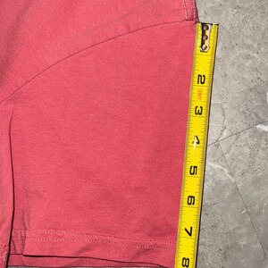 May include: A section of a coral-colored t-shirt is shown with a yellow measuring tape. The tape shows the shirt's length is approximately 8 inches. The shirt's hem is visible, with a double-stitched seam.