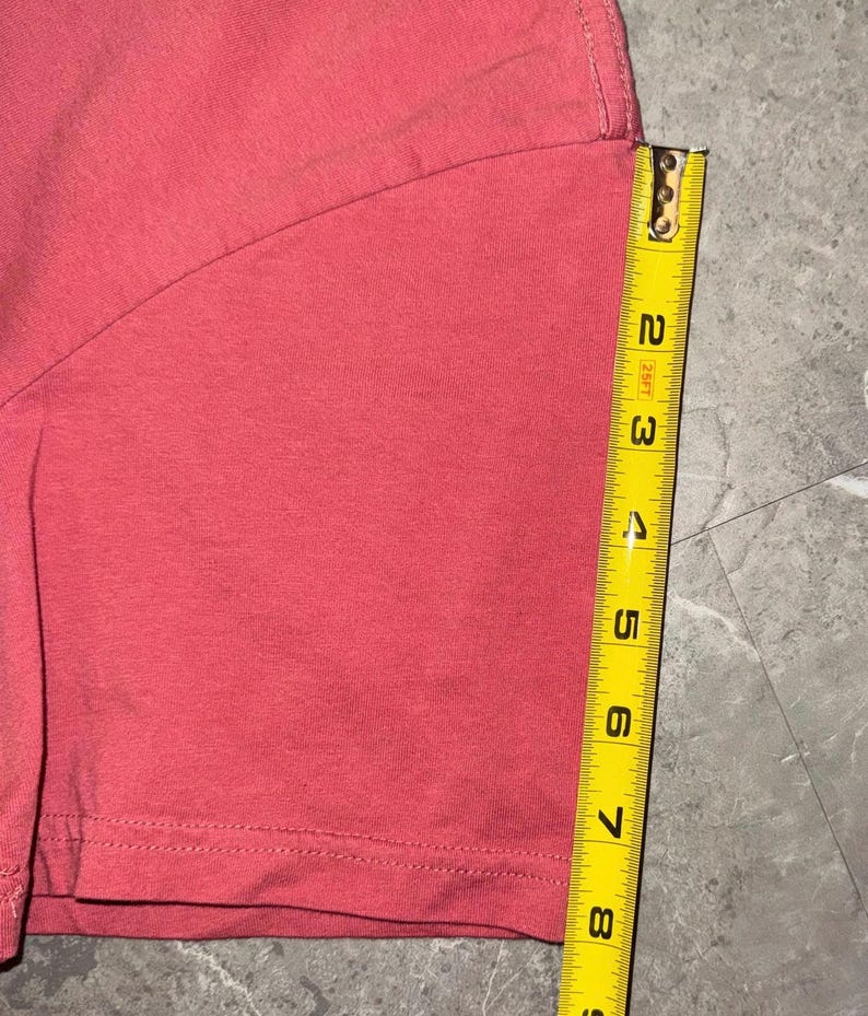 May include: A section of a coral-colored t-shirt is shown with a yellow measuring tape. The tape shows the shirt's length is approximately 8 inches. The shirt's hem is visible, with a double-stitched seam.