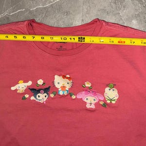 May include: A red XL t-shirt featuring embroidered cartoon characters. The shirt has a crew neck and short sleeves. The characters include Hello Kitty and other Sanrio characters. The shirt measures approximately 21 inches across.