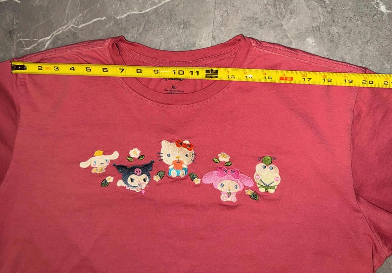 May include: A red XL t-shirt featuring embroidered cartoon characters. The shirt has a crew neck and short sleeves. The characters include Hello Kitty and other Sanrio characters. The shirt measures approximately 21 inches across.
