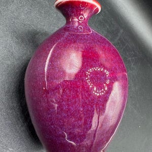 Puede incluir: Un pequeño jarrón decorativo con un esmalte brillante de color púrpura intenso. El jarrón tiene un cuerpo redondeado, un cuello estrecho y un borde acampanado. El borde es rojo con un acento blanco. La base es blanca. El jarrón es probablemente de cerámica.