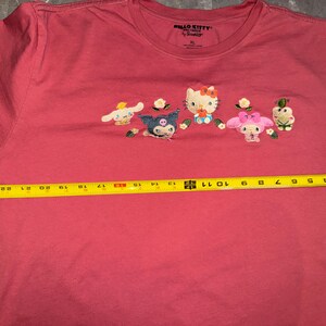 May include: A coral-colored t-shirt featuring embroidered Hello Kitty characters. The shirt, size XL, has a crew neck and short sleeves. The characters include Hello Kitty, My Melody, and others. The shirt's width is approximately 11 inches.