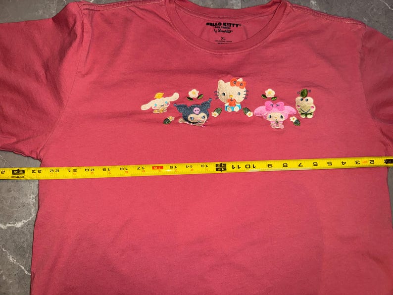 May include: A coral-colored t-shirt featuring embroidered Hello Kitty characters. The shirt, size XL, has a crew neck and short sleeves. The characters include Hello Kitty, My Melody, and others. The shirt's width is approximately 11 inches.