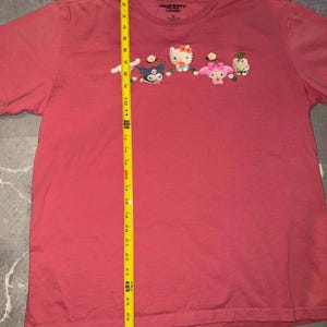 May include: A red XL Hello Kitty t-shirt featuring embroidered Hello Kitty characters. The shirt has a crew neck and short sleeves. The characters include Hello Kitty, My Melody, and other Sanrio characters.