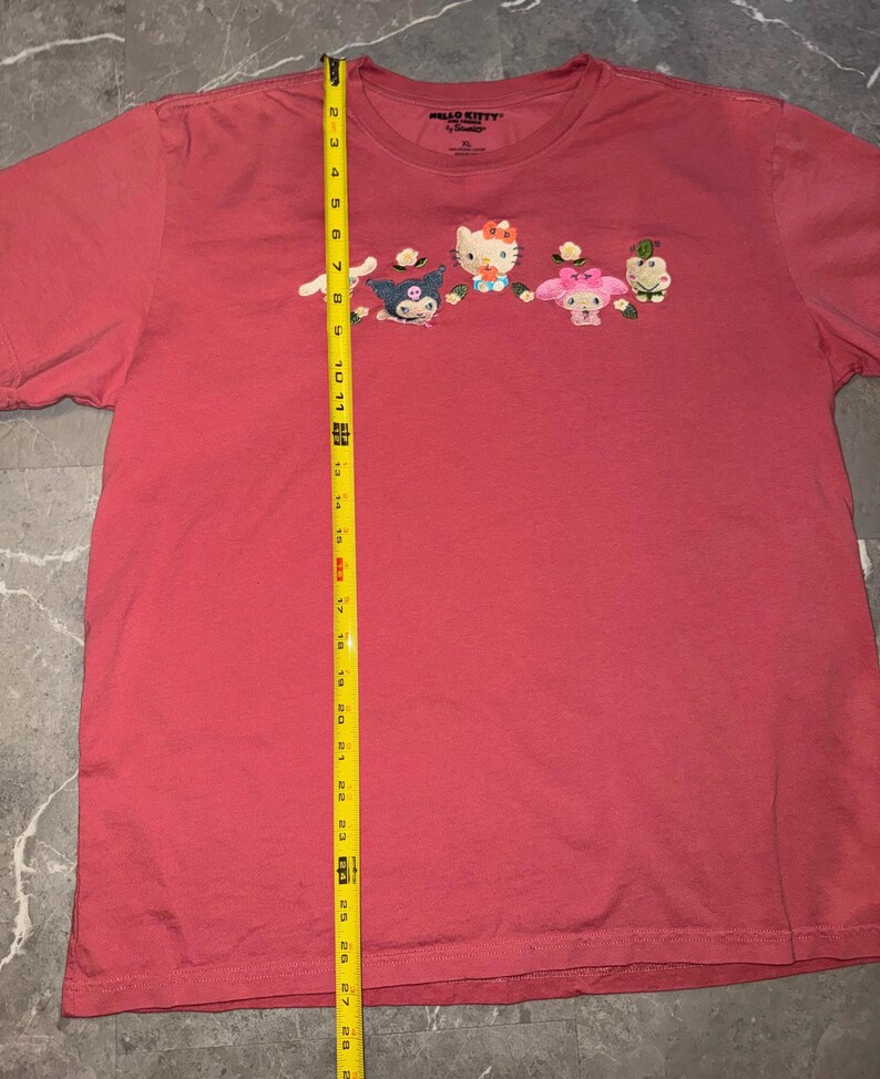 May include: A red XL Hello Kitty t-shirt featuring embroidered Hello Kitty characters. The shirt has a crew neck and short sleeves. The characters include Hello Kitty, My Melody, and other Sanrio characters.