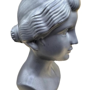 Puede incluir: Busto plateado de una mujer, visto de perfil. El peinado es un mo&ntilde;o. La escultura muestra los rasgos faciales y el cuello. El busto est&aacute; hecho de un material liso y es un objeto decorativo.