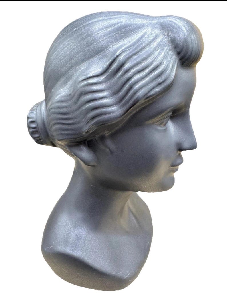 Puede incluir: Busto plateado de una mujer, visto de perfil. El peinado es un mo&ntilde;o. La escultura muestra los rasgos faciales y el cuello. El busto est&aacute; hecho de un material liso y es un objeto decorativo.