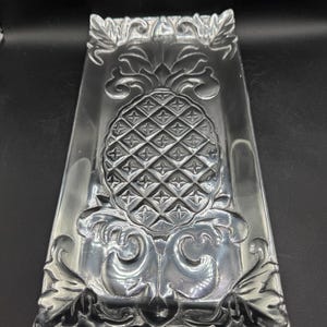 May include: A rectangular silver-toned metal tray featuring a central embossed pineapple motif. The tray has decorative scrollwork and leaf-like patterns along the edges and surrounding the pineapple design, with a polished, reflective surface finish.