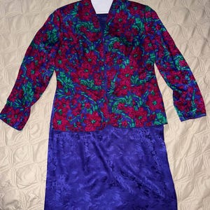 Peut inclure: Ensemble deux pi&egrave;ces vintage avec une veste &agrave; imprim&eacute; floral et une jupe violette unie. La veste pr&eacute;sente un motif vibrant de fleurs rouges, de feuilles vertes et un fond bleu. La jupe est violette unie avec un motif floral subtil.