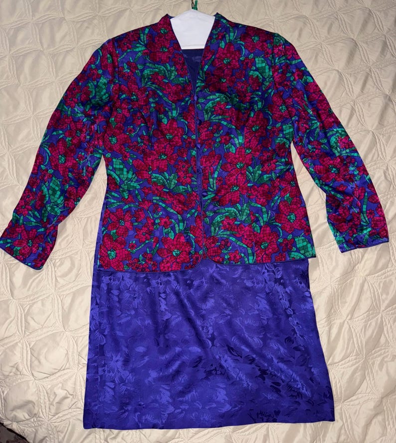 Peut inclure: Ensemble deux pi&egrave;ces vintage avec une veste &agrave; imprim&eacute; floral et une jupe violette unie. La veste pr&eacute;sente un motif vibrant de fleurs rouges, de feuilles vertes et un fond bleu. La jupe est violette unie avec un motif floral subtil.
