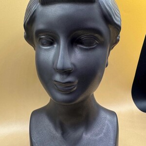 Puede incluir: Busto de cer&aacute;mica negra mate de la cabeza y los hombros de una mujer. La escultura presenta rasgos faciales detallados, incluyendo ojos cerrados, una nariz definida y una suave sonrisa. El cabello est&aacute; peinado en un mo&ntilde;o cl&aacute;sico.