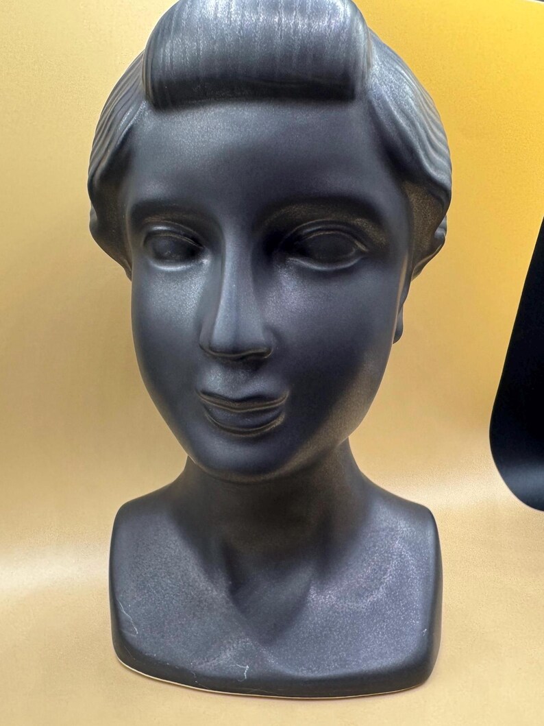 Puede incluir: Busto de cer&aacute;mica negra mate de la cabeza y los hombros de una mujer. La escultura presenta rasgos faciales detallados, incluyendo ojos cerrados, una nariz definida y una suave sonrisa. El cabello est&aacute; peinado en un mo&ntilde;o cl&aacute;sico.