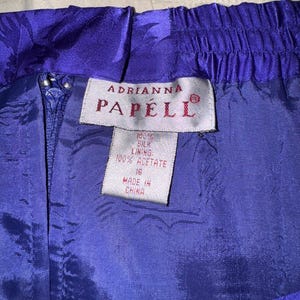Peut inclure: Gros plan sur une jupe violette vibrante avec un motif floral. La jupe est dot&eacute;e d'une fermeture &eacute;clair et d'une taille &eacute;lastique. L'&eacute;tiquette indique "Adrianna Papell" et indique les mati&egrave;res : doublure 100% soie et 100% ac&eacute;tate. Taille 16, fabriqu&eacute; en Chine.