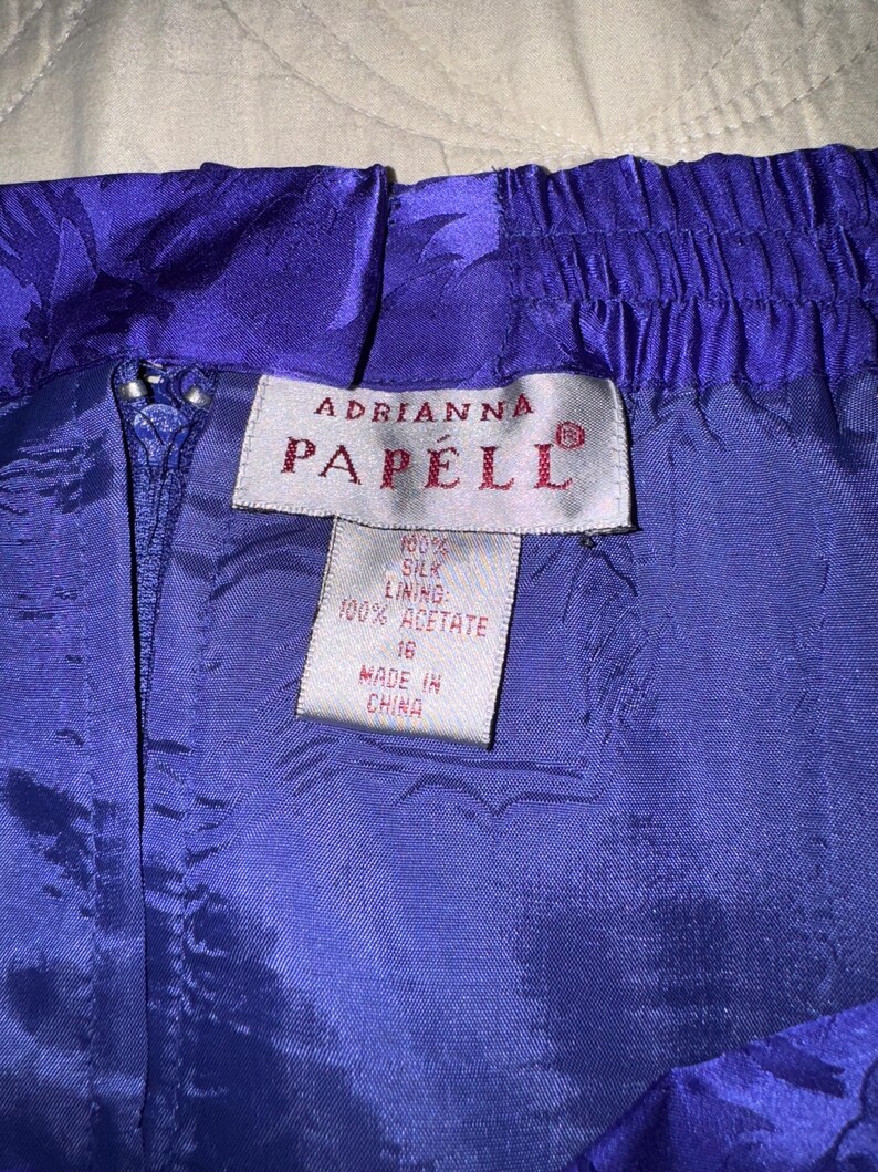 Peut inclure: Gros plan sur une jupe violette vibrante avec un motif floral. La jupe est dot&eacute;e d'une fermeture &eacute;clair et d'une taille &eacute;lastique. L'&eacute;tiquette indique "Adrianna Papell" et indique les mati&egrave;res : doublure 100% soie et 100% ac&eacute;tate. Taille 16, fabriqu&eacute; en Chine.