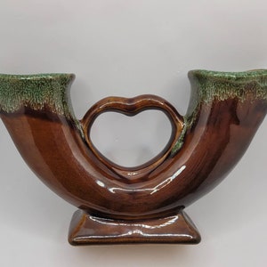 Puede incluir: Un jarrón de cerámica marrón con un esmalte verde. El jarrón tiene dos aberturas y un asa en forma de corazón.