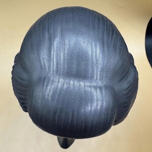 Puede incluir: Busto estilizado gris oscuro de una persona con un mo&ntilde;o. La escultura tiene un acabado mate y liso, vista desde atr&aacute;s, mostrando la textura detallada del cabello y la forma de la cabeza.
