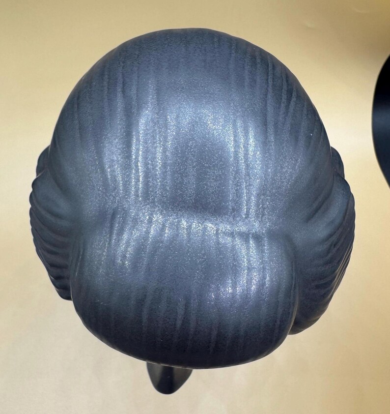 Puede incluir: Busto estilizado gris oscuro de una persona con un mo&ntilde;o. La escultura tiene un acabado mate y liso, vista desde atr&aacute;s, mostrando la textura detallada del cabello y la forma de la cabeza.