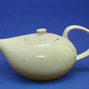 May include: A pale cream-coloured ceramic teapot with a smooth, rounded body, a short spout, a matching lid with a small knob handle, and a curved side handle. The teapot features a glossy finish and a wide, low-profile design for serving tea.