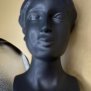 Puede incluir: Busto negro mate de una mujer con pelo corto y expresi&oacute;n serena. La escultura tiene un acabado liso y est&aacute; sobre un fondo neutro. El busto es un objeto decorativo.