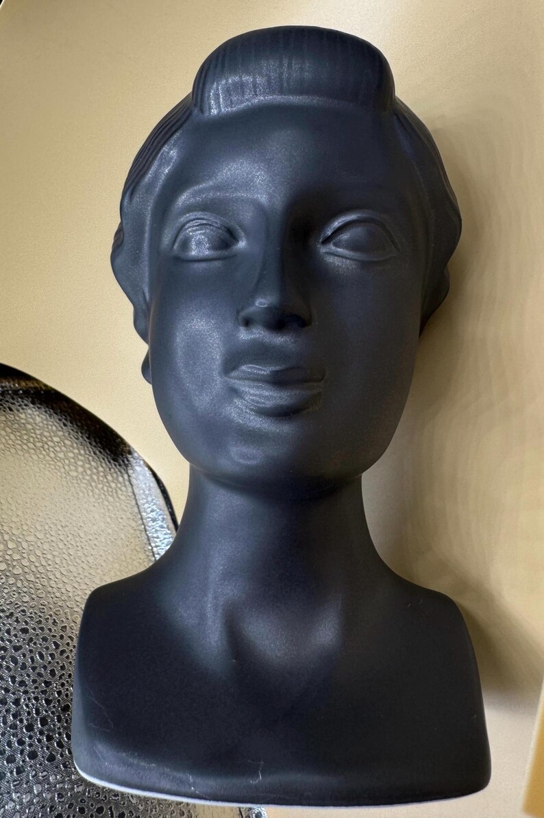 Puede incluir: Busto negro mate de una mujer con pelo corto y expresi&oacute;n serena. La escultura tiene un acabado liso y est&aacute; sobre un fondo neutro. El busto es un objeto decorativo.