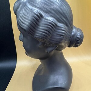 Puede incluir: Busto gris oscuro de la cabeza y los hombros de una mujer, esculpido en estilo cl&aacute;sico. El cabello est&aacute; peinado en un mo&ntilde;o en la parte posterior de la cabeza. El busto tiene un acabado liso y mate.