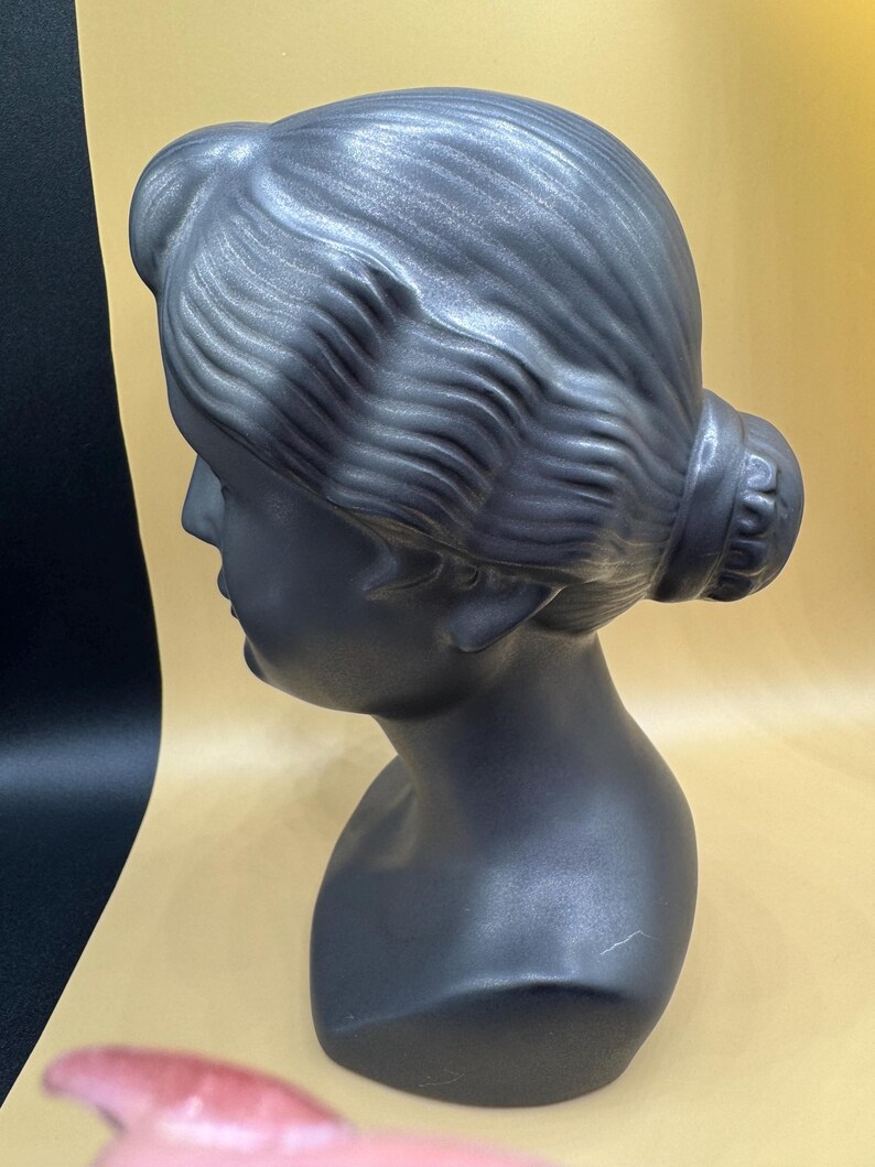 Puede incluir: Busto gris oscuro de la cabeza y los hombros de una mujer, esculpido en estilo cl&aacute;sico. El cabello est&aacute; peinado en un mo&ntilde;o en la parte posterior de la cabeza. El busto tiene un acabado liso y mate.