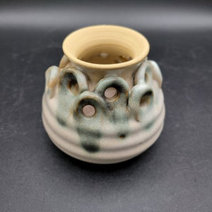 May include: A small, ceramic pot with a light brown glaze and green accents. The pot has a unique design with a series of holes around the top and a textured, wavy pattern around the bottom.