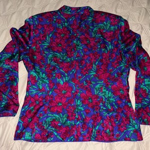Peut inclure: Un chemisier &agrave; manches longues avec un motif floral vibrant. Le chemisier pr&eacute;sente un fond bleu riche orn&eacute; de grandes fleurs magenta et de feuillage vert. Le motif se r&eacute;p&egrave;te sur tout le v&ecirc;tement.
