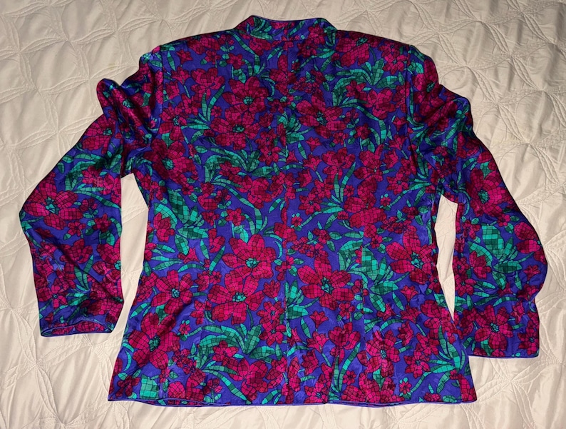 Peut inclure: Un chemisier &agrave; manches longues avec un motif floral vibrant. Le chemisier pr&eacute;sente un fond bleu riche orn&eacute; de grandes fleurs magenta et de feuillage vert. Le motif se r&eacute;p&egrave;te sur tout le v&ecirc;tement.