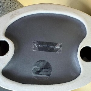 Puede incluir: Una bandeja decorativa de cer&aacute;mica con interior gris y borde blanco irregular. La bandeja tiene dos hendiduras circulares negras y una etiqueta que dice "by ANTHROPOLOGIE".