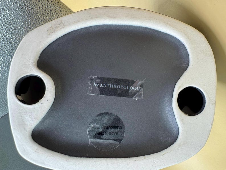 Puede incluir: Una bandeja decorativa de cer&aacute;mica con interior gris y borde blanco irregular. La bandeja tiene dos hendiduras circulares negras y una etiqueta que dice "by ANTHROPOLOGIE".