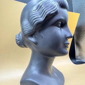 Puede incluir: Busto de cer&aacute;mica gris oscuro de una mujer con detalles en el cabello y rasgos faciales. El busto es un perfil, mostrando las elegantes l&iacute;neas de la cara y el cuello. La base es blanca.