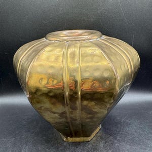 May include: A metallic gold-toned vase with a wide, rounded body and a narrow neck. The surface features a repeating vertical panel design with a subtle hammered texture throughout, creating a reflective finish with varied light and dark highlights.