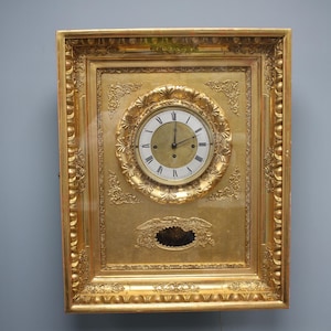 Puede incluir: Reloj de pared dorado ornamentado con marco rectangular. La esfera del reloj es blanca con números romanos negros. El marco presenta intrincadas tallas y un óvalo decorativo alrededor de la esfera. El reloj mide aproximadamente 46 cm de altura.