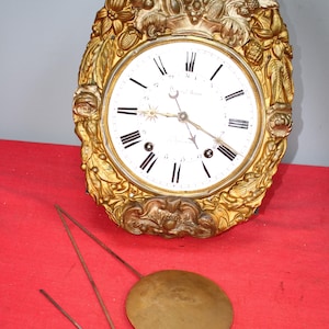 May include: A gold-colored, ornate clock with a white face and black Roman numeral hour markers. The clock is surrounded by a decorative frame with floral and fruit motifs. The clock is hanging on a red wall and has a pendulum hanging below it.