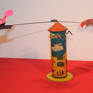 May include: A vintage tin toy with a yellow base and a blue and yellow tower. The tower has a red and white airplane and a white airplane painted on it. Two red and white airplanes are attached to the tower by a metal rod and spin around.
