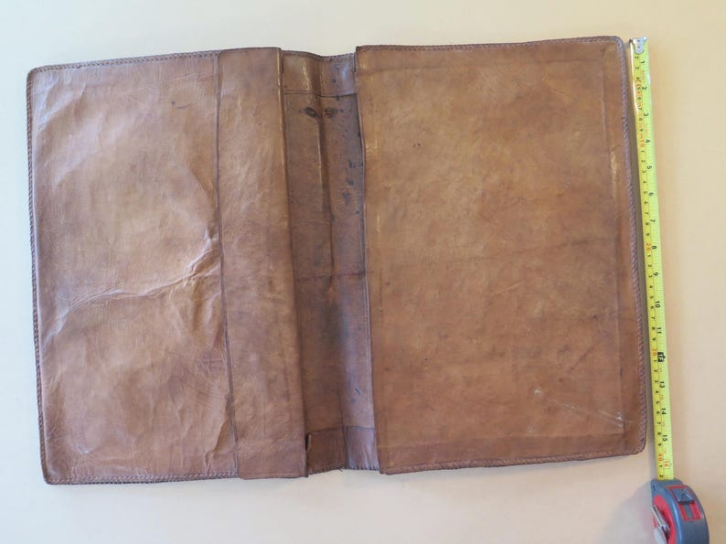 K&ouml;nnte beinhalten: Offenes, braunes Lederjournal mit abgenutztem Aussehen. Das Journal ist ge&ouml;ffnet und zeigt leere Seiten. Ein gelbes Ma&szlig;band liegt neben dem Journal und zeigt eine L&auml;nge von etwa 30 cm.