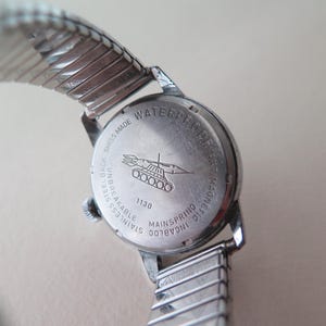 Op de afbeelding: Een zilveren horloge met een roestvrijstalen achterkant. De achterkant van het horloge heeft de tekst "WATERPROOF 5 ATM MAGNETIC", "STAINLESS STEEL BACK UNBREAKABLE", "SWISS MADE" en "INCABLOC MAINSPRING 1130". Het horloge heeft ook een klein beeld van een tank met een raket erop.