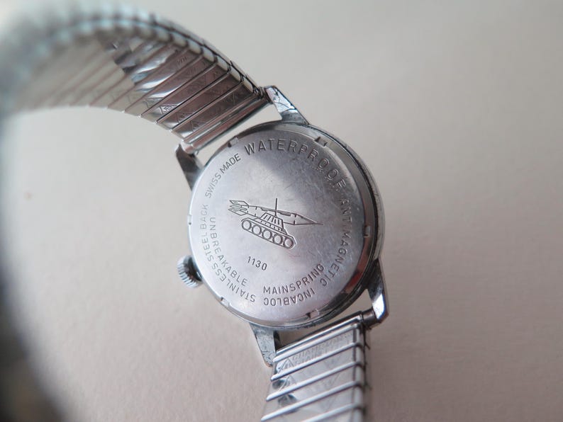 Op de afbeelding: Een zilveren horloge met een roestvrijstalen achterkant. De achterkant van het horloge heeft de tekst "WATERPROOF 5 ATM MAGNETIC", "STAINLESS STEEL BACK UNBREAKABLE", "SWISS MADE" en "INCABLOC MAINSPRING 1130". Het horloge heeft ook een klein beeld van een tank met een raket erop.