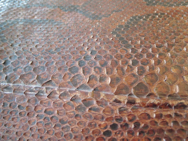 K&ouml;nnte beinhalten: Nahaufnahme einer strukturierten Oberfl&auml;che mit einem sich wiederholenden Muster aus braunen und schwarzen Schuppen. Die Schuppen haben eine erhabene, unebene Textur, und der Gesamteffekt erinnert an Reptilienhaut.