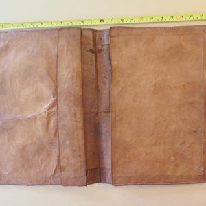 K&ouml;nnte beinhalten: Offenes, braunes Lederjournal mit einer gen&auml;hten Bord&uuml;re. Das Journal ist im ge&ouml;ffneten Zustand etwa 61 cm lang und 41 cm breit. Das Leder hat ein abgenutztes, strukturiertes Aussehen mit sichtbaren Falten und Farbvariationen.