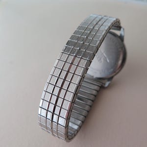 Op de afbeelding: Een zilveren horloge met een metalen band. De wijzerplaat van het horloge is niet zichtbaar. De band is van een metalen gaas.