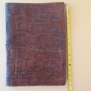 K&ouml;nnte beinhalten: Eine braune Ledermappe mit Schlangenmuster. Die Mappe ist rechteckig und etwa 40 cm lang. Ein gelbes Ma&szlig;band liegt neben der Mappe.