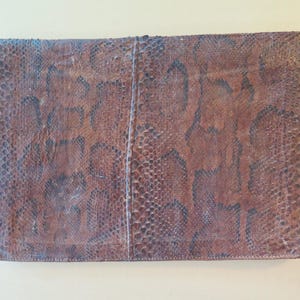 Könnte beinhalten: Eine rechteckige braune Clutch mit Schlangenmuster. Die Clutch hat einen Klappenverschluss und eine strukturierte Oberfläche. Die Farbe ist eine Mischung aus Braun und Dunkelbraun mit einem dezenten Glanz. Das Gesamtdesign ist schlicht und elegant.