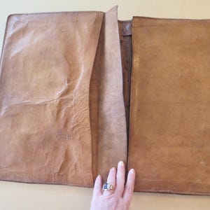 K&ouml;nnte beinhalten: Offenes, hellbraunes Leder-Portfolio mit strukturierter Oberfl&auml;che. Das Portfolio ist ge&ouml;ffnet und zeigt eine Tasche und das Innere. Die Kanten sind mit einem dunkleren Faden gen&auml;ht. Das Portfolio ist rechteckig und scheint handgefertigt zu sein.