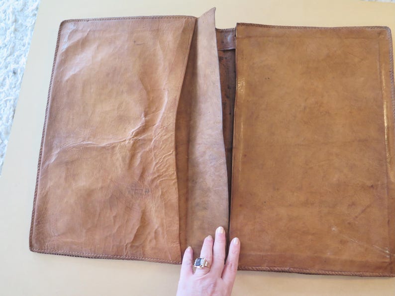 K&ouml;nnte beinhalten: Offenes, hellbraunes Leder-Portfolio mit strukturierter Oberfl&auml;che. Das Portfolio ist ge&ouml;ffnet und zeigt eine Tasche und das Innere. Die Kanten sind mit einem dunkleren Faden gen&auml;ht. Das Portfolio ist rechteckig und scheint handgefertigt zu sein.