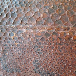 K&ouml;nnte beinhalten: Nahaufnahme einer strukturierten Oberfl&auml;che mit einem Muster, das an Reptilienhaut erinnert. Die Farbpalette umfasst Braunt&ouml;ne, Schwarz und Beige, wodurch eine nat&uuml;rliche, organische &Auml;sthetik entsteht. Die Oberfl&auml;che scheint aus einem haltbaren Material zu bestehen.