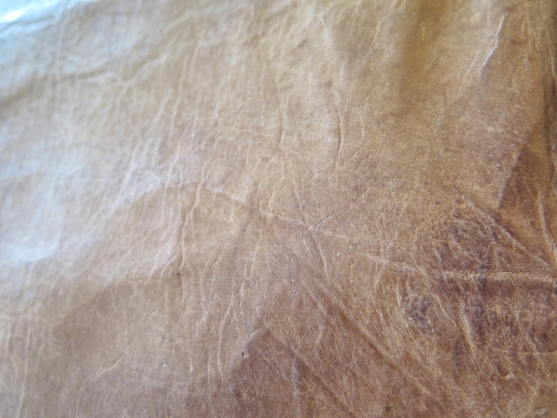 K&ouml;nnte beinhalten: Nahaufnahme einer strukturierten, hellbraunen Lederoberfl&auml;che. Das Leder hat ein abgenutztes Aussehen mit sichtbaren Falten und Farbvariationen, was eine rustikale &Auml;sthetik erzeugt. Die Oberfl&auml;che ist beleuchtet und hebt die nat&uuml;rliche Maserung und Unvollkommenheiten hervor.